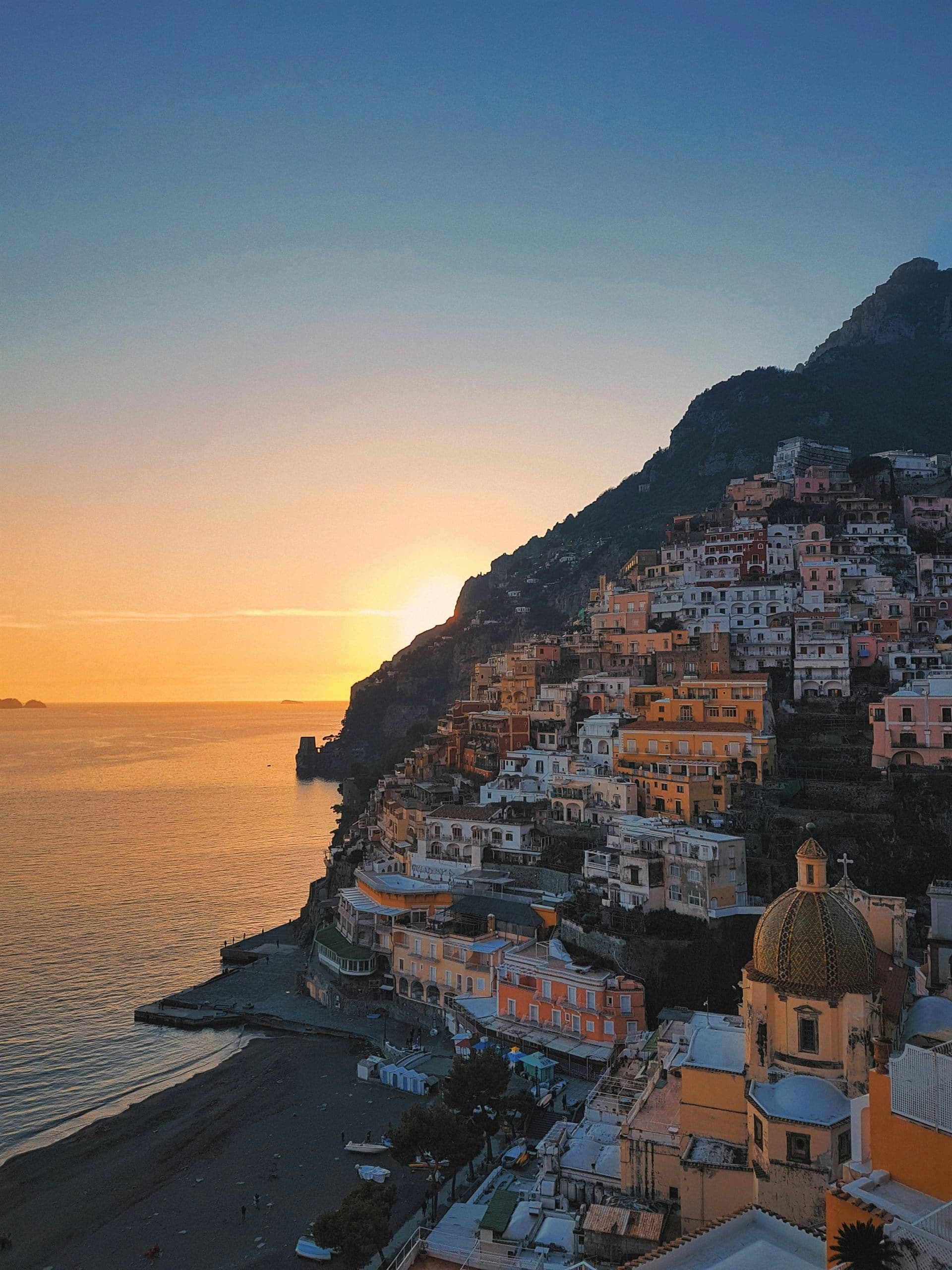Pastel houses stacked up a steep cliff above a beach and harbour on the Amalfi Coast at sunset, a tiled church dome in the foreground.