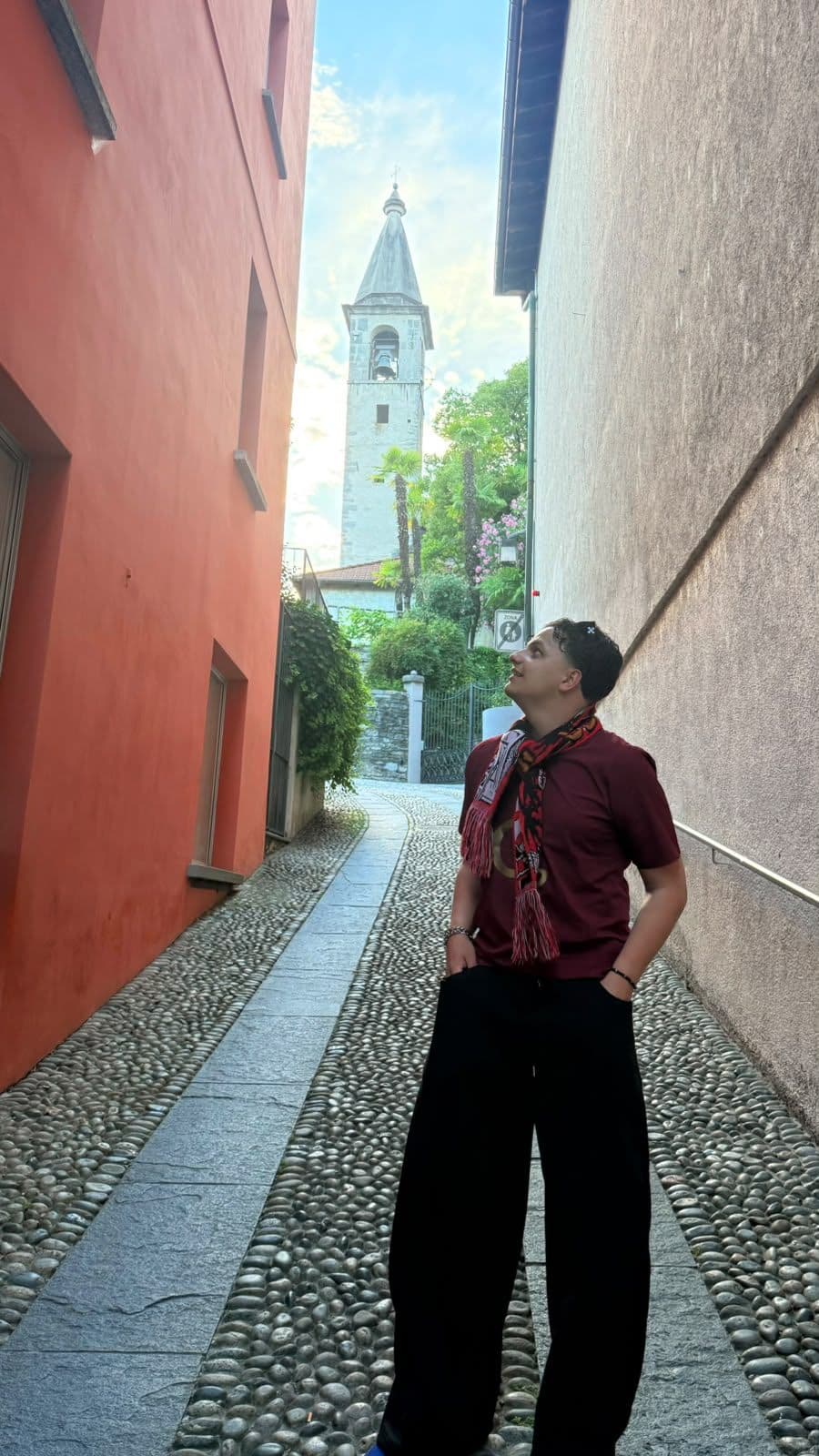 A person standing in a narrow stone-paved lane, looking up towards a bell tower rising above the rooftops.