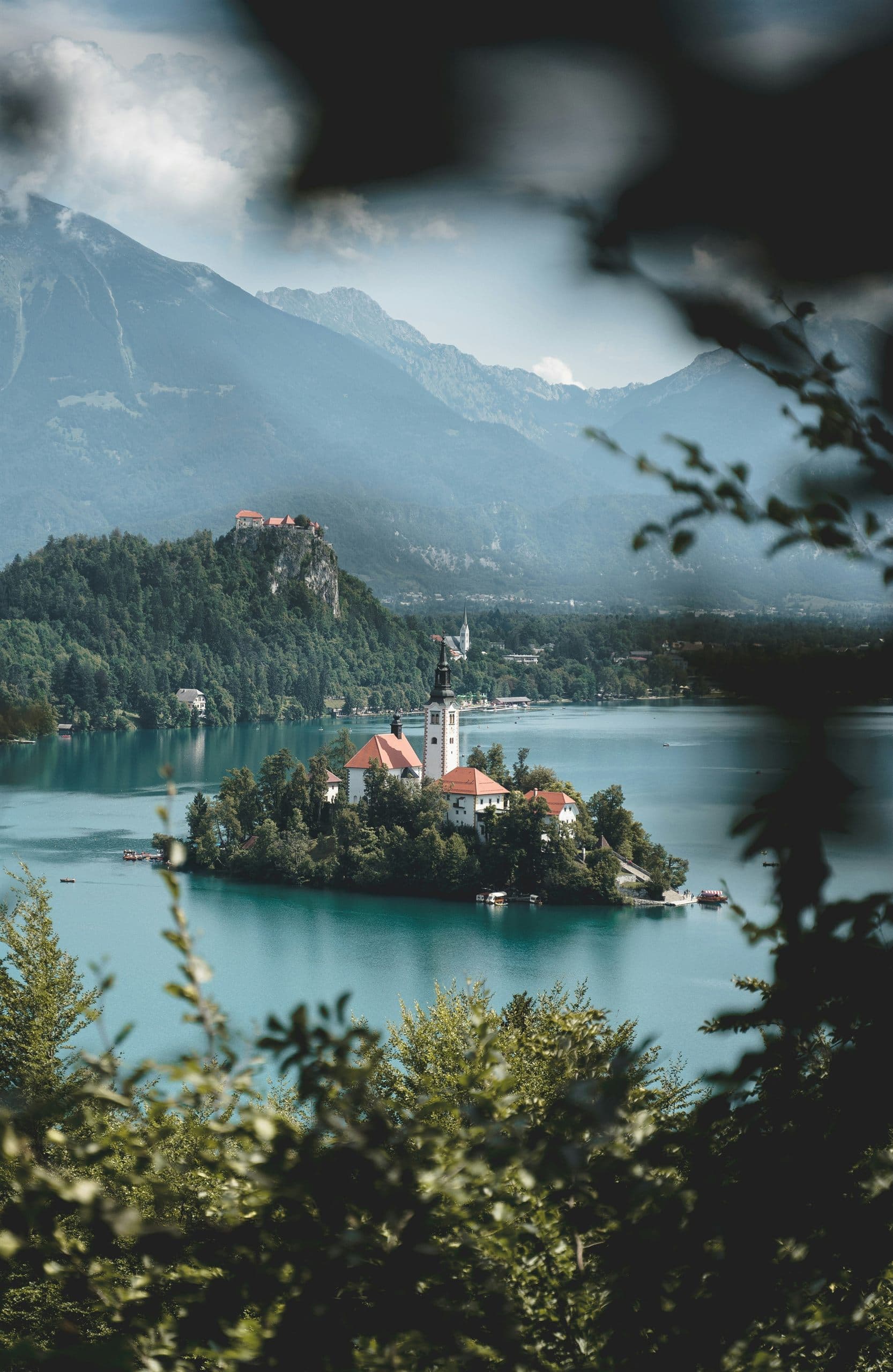 A small wooded island with a church and bell tower on a turquoise mountain lake, forested slopes and high peaks behind it, framed by leaves in the foreground.