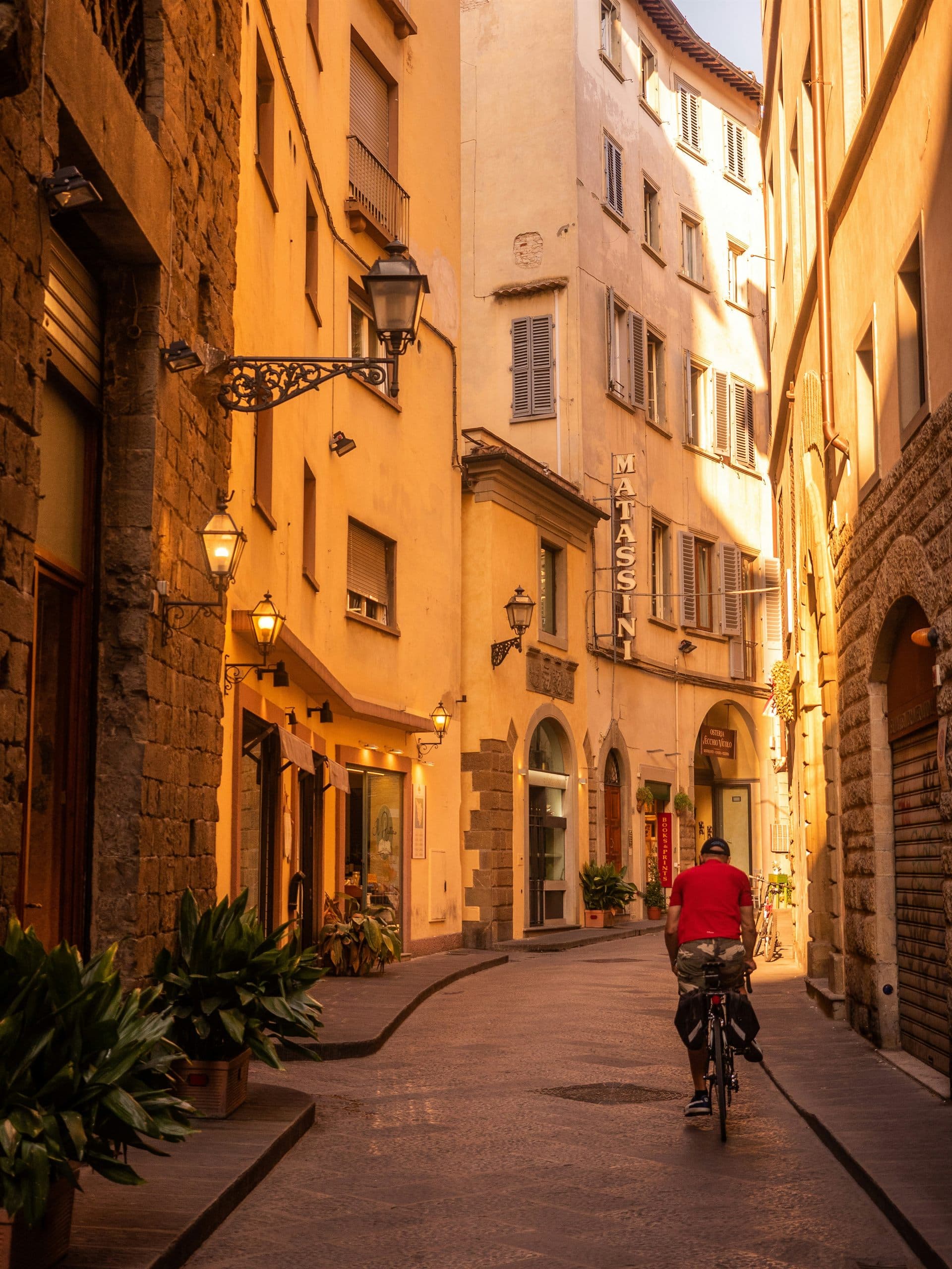A cyclist riding down a narrow street of stone buildings and iron lanterns in low evening sun.