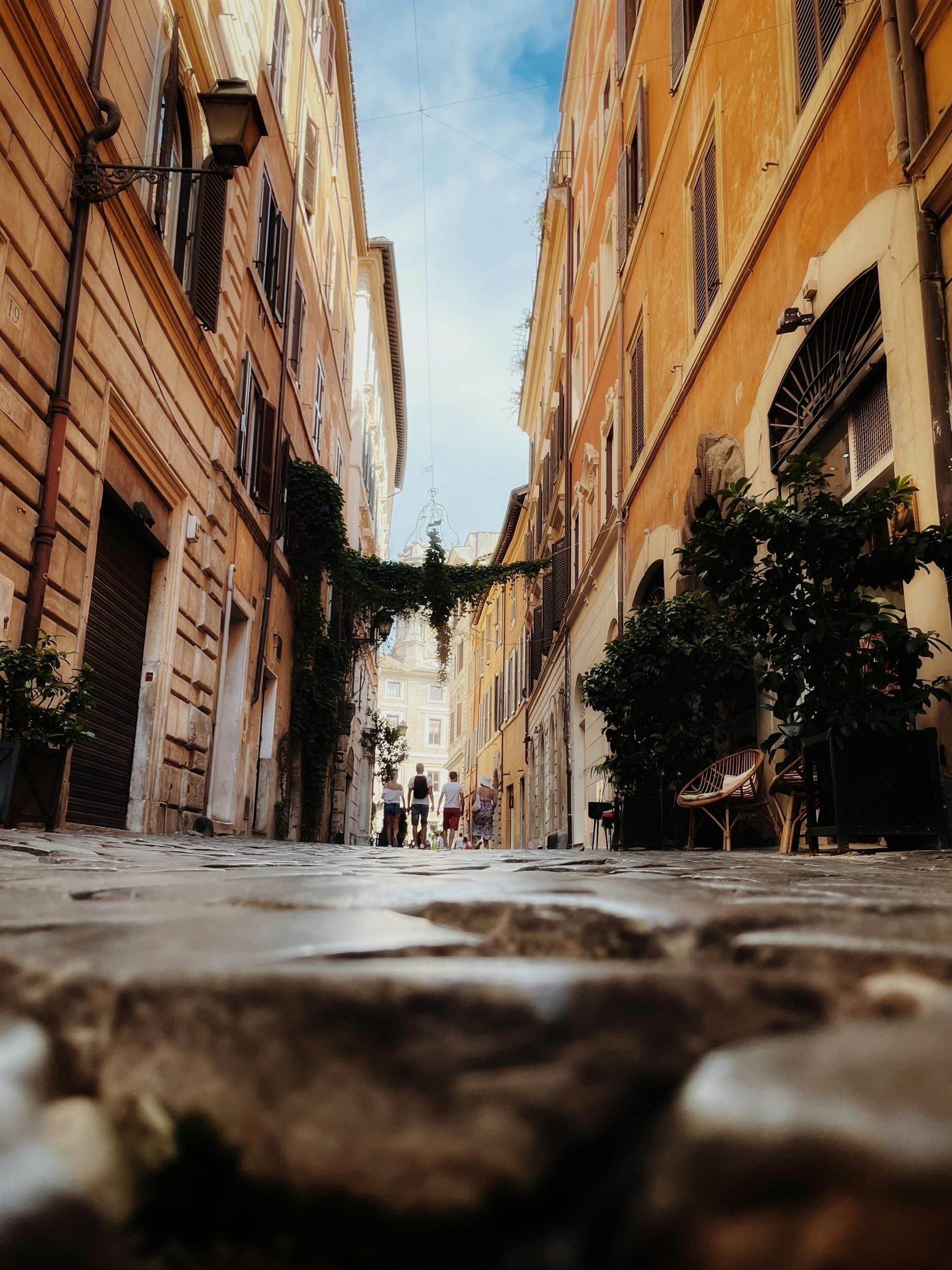 A narrow cobbled street between ochre and terracotta buildings, with people walking towards a church facade at the far end.