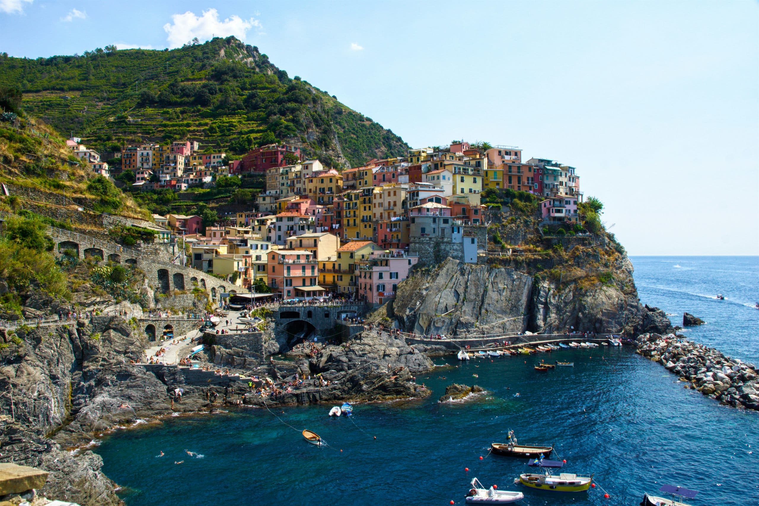 A village of pastel houses stacked on a rocky headland above a small harbour, with terraced hillsides rising behind it and boats drawn up on the slipway below.