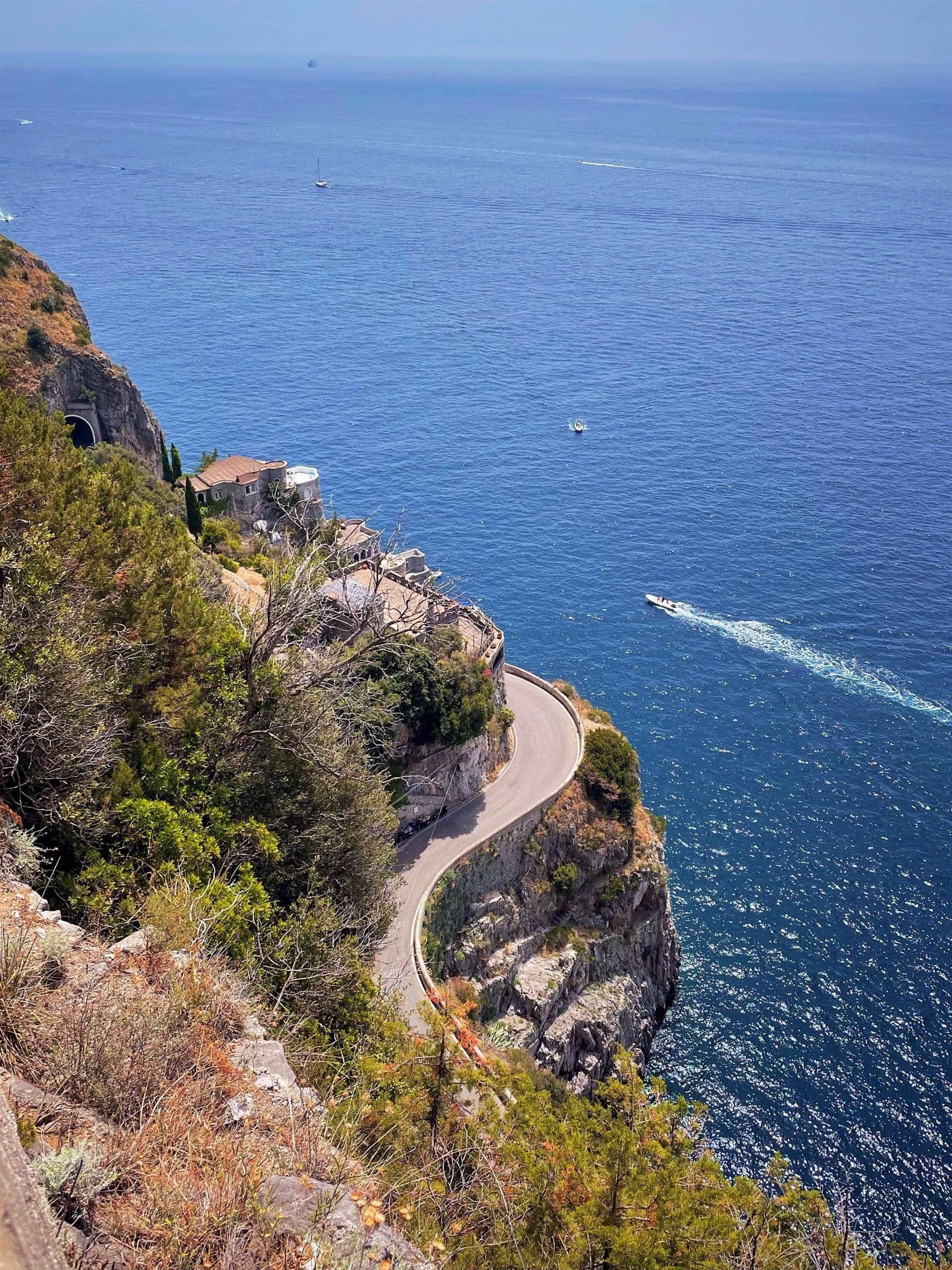 A narrow coast road curving around a cliff high above deep blue sea, with a boat passing below.