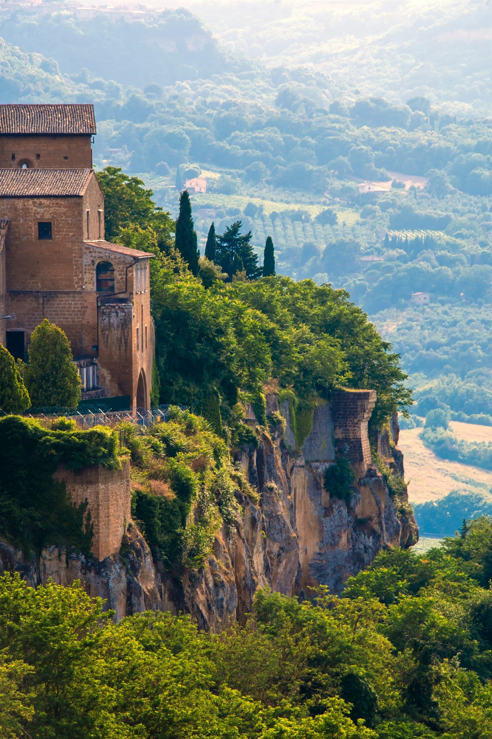 A stone building standing at the edge of a steep tufa cliff above a green valley of fields and trees.