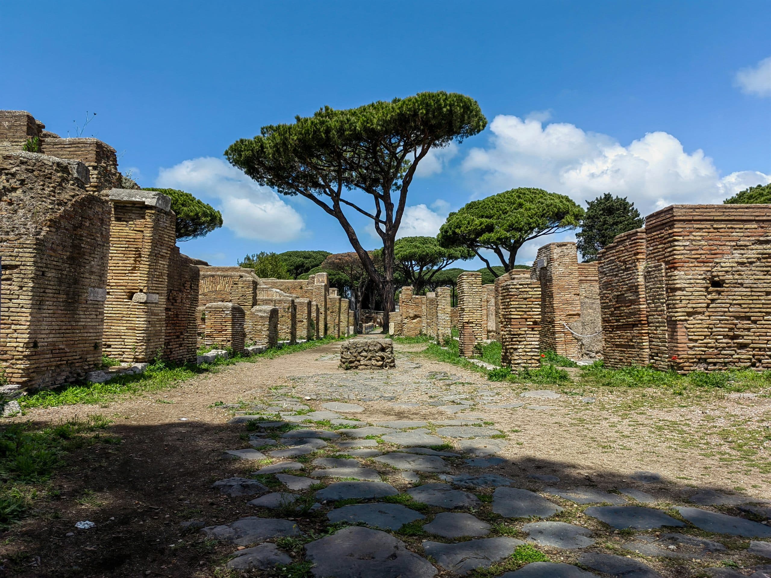 A paved Roman road running between low brick ruins and umbrella pines under a blue sky.