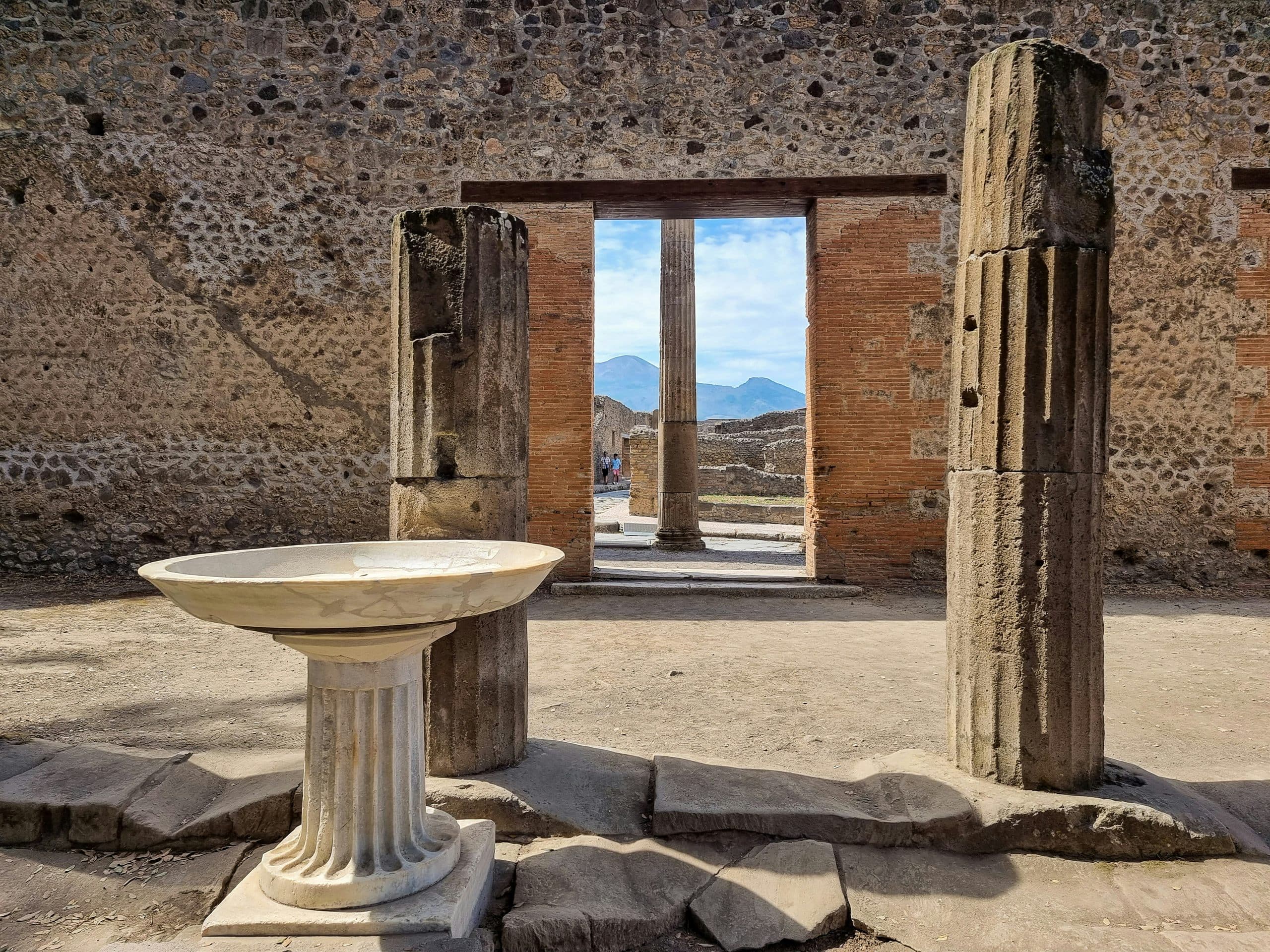 A marble basin and broken stone columns in a walled courtyard at Pompeii, with Vesuvius framed in the doorway beyond.
