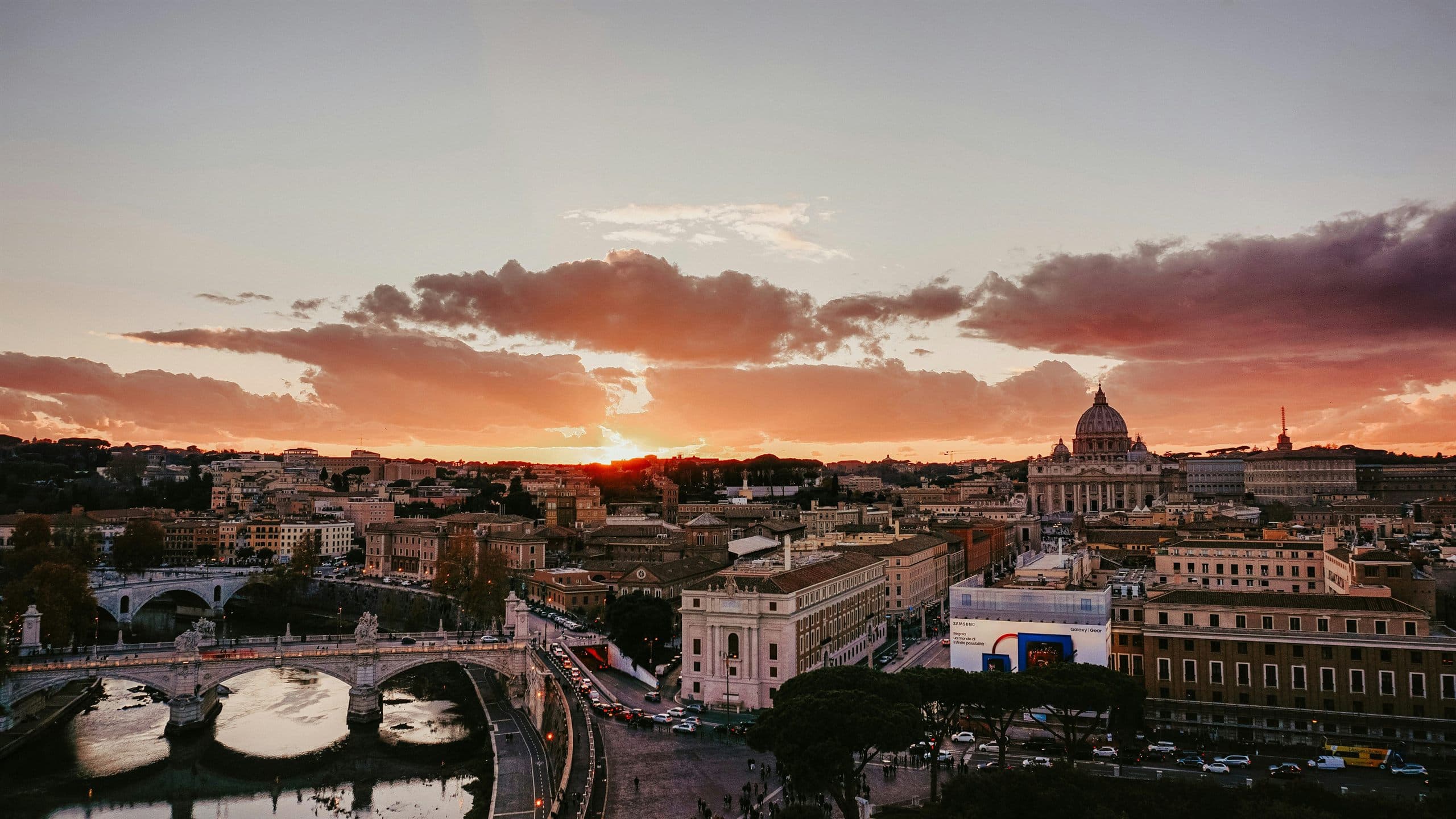 Rome at sunset seen from above, the dome of St Peter's Basilica on the skyline and a bridge crossing the Tiber below.