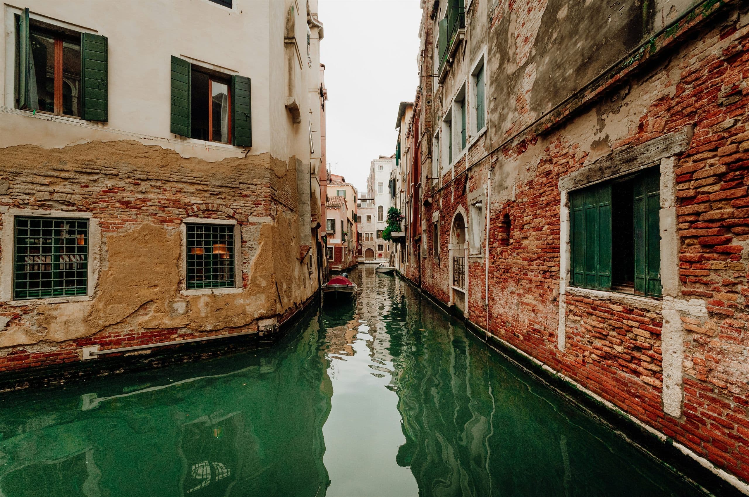 A narrow Venetian canal between brick and stucco walls, with green shutters, a small moored boat and a low water door at the waterline.
